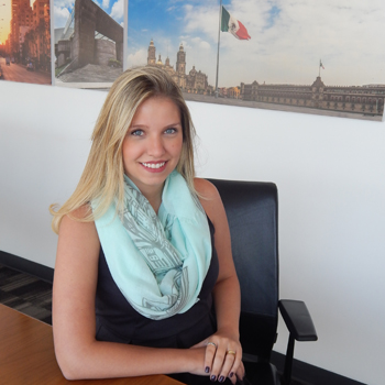 Persona sentada junto a una mesa, con fotografías de una ciudad al fondo.