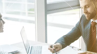 Dos personas conversan en una oficina con una laptop abierta.