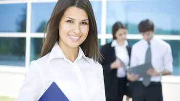 Persona sonriente con una carpeta azul, con dos personas trabajando atrás.