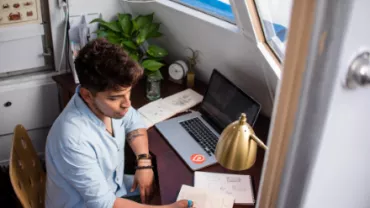 Persona dibujando en un cuaderno en un escritorio con laptop y planta.