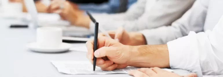Personas escribiendo en una reunión, con tazas y computadoras portátiles en una mesa.