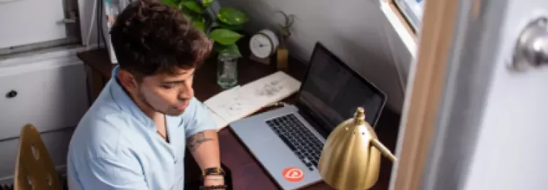 Persona dibujando en un cuaderno en un escritorio con laptop y planta.