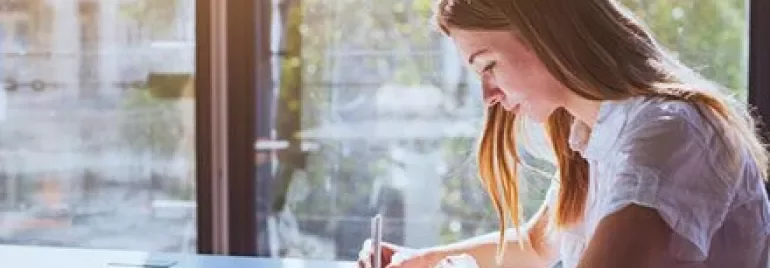 Persona escribiendo en un cuaderno cerca de una ventana soleada.