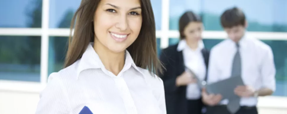 Persona sonriente con una carpeta azul, con dos personas trabajando atrás.